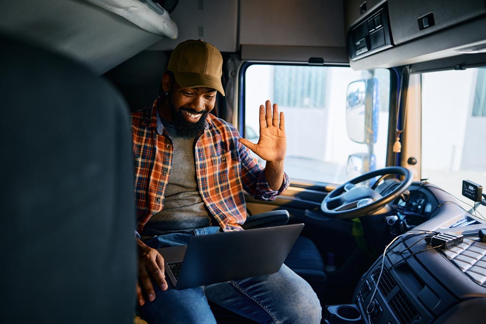 Happy African American driver waving during video call over laptop on truck parking lot.